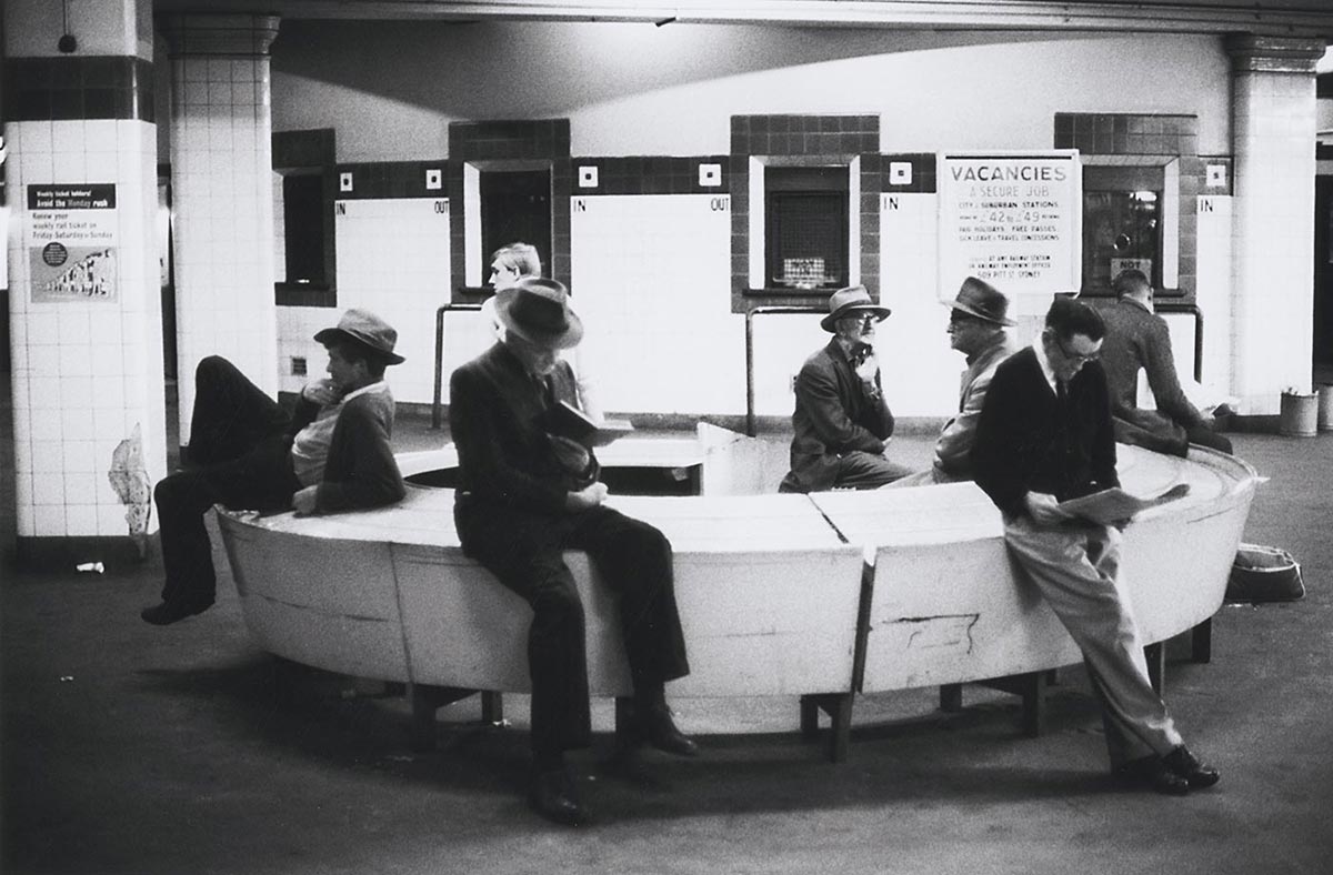 Interior photo of seven men sitting on a circular bench cum table. A few of them are reading but most are doing nothing at all. In the background are the station’s ticket booths. Next to one of these is a poster advertising railway jobs.