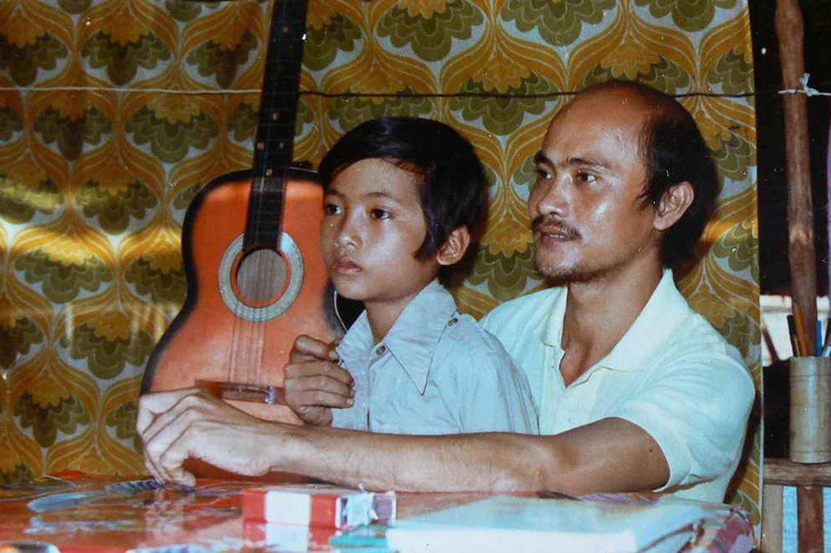 A man sits at a table with a young boy on his lap. Behind them a guitar is visible. - click to view larger image