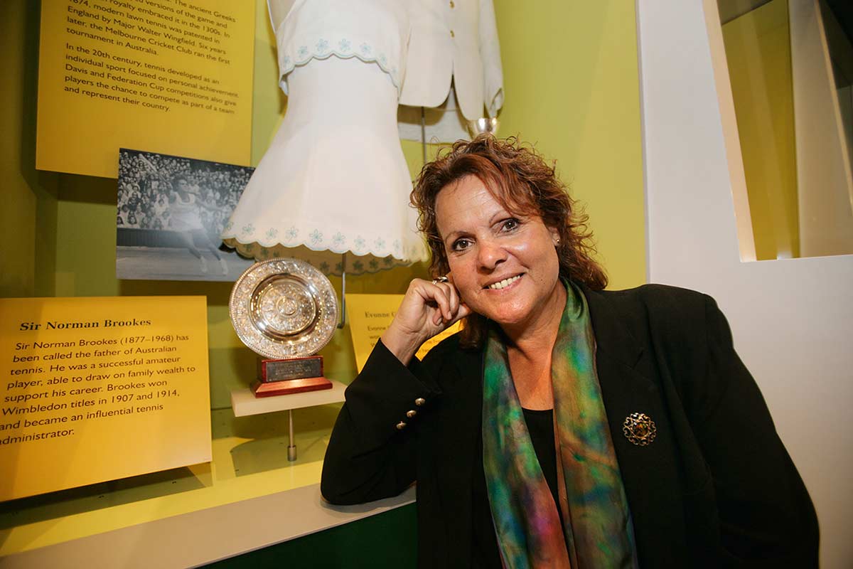 Evonne Goolagong Cawley sits beside a case containing a small circular trophy, white tennis dress, text panels and black and white photograph. - click to view larger image