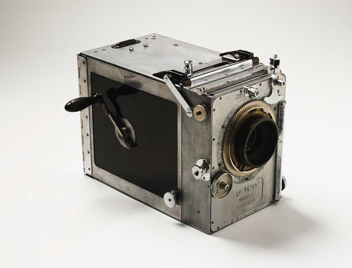 A rectangular box-shaped motion picture camera with a hand-crank and glass lens. The camera's metal body is silver with black side panels and has external slides, levers and dials for adjustment and operation. 'Le Parvo Modele L' is stamped in the metal plate beneath the lens at the front of the camera.