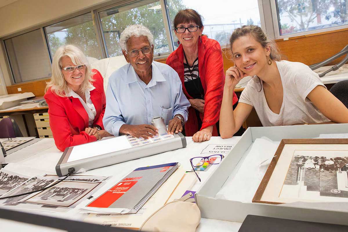 A man and three women standing at a bench, with photos and a lightbox on the benchtop. - click to view larger image