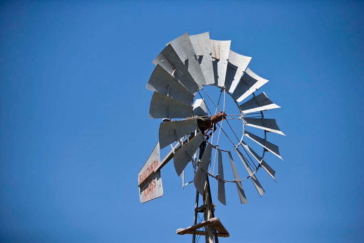 Top section of a windmill, set against a blue sky. Grey blades in a circular formation front a sail with 'SOUTHERN CROSS' painted in red. - click to view larger image