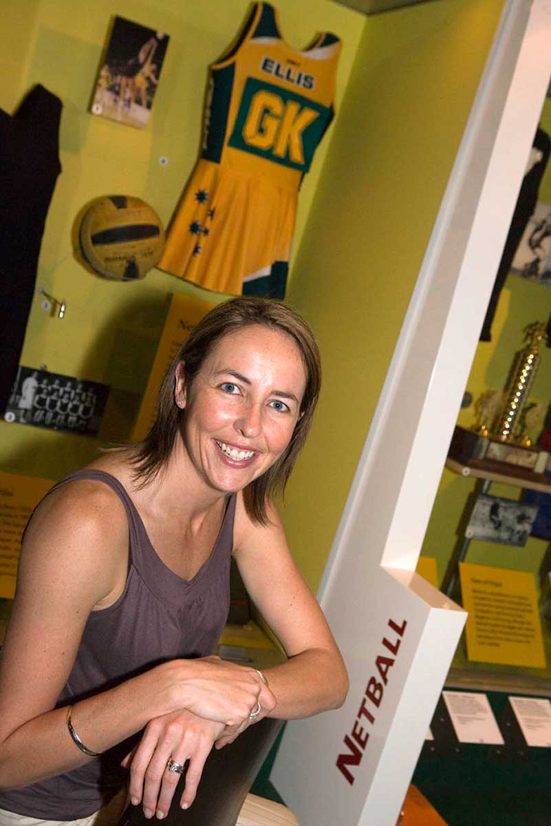 Liz Ellis sits in front of display case showing images, netball and green and gold 'Ellis GK' uniform. - click to view larger image