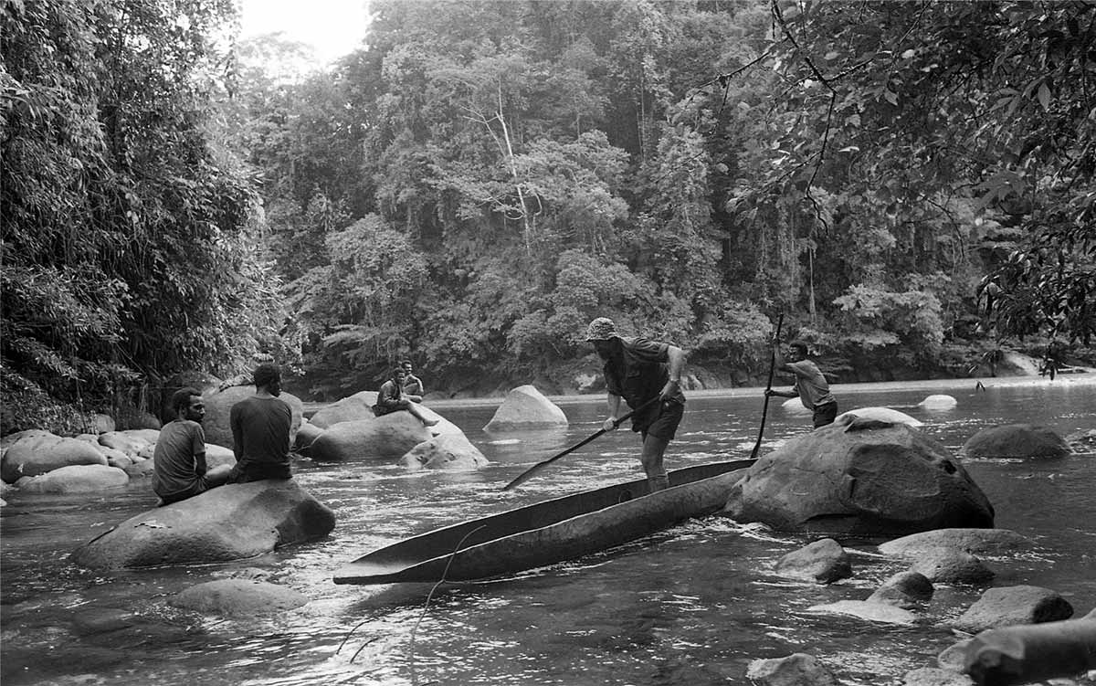 Black and white photo on men in canoe with others seated on rocks in Purari River.