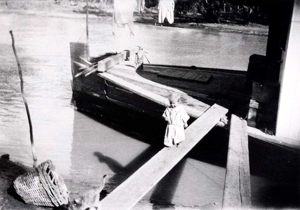 Black and white photo of a young child walking along a gangplank, following a cat. - click to view larger image