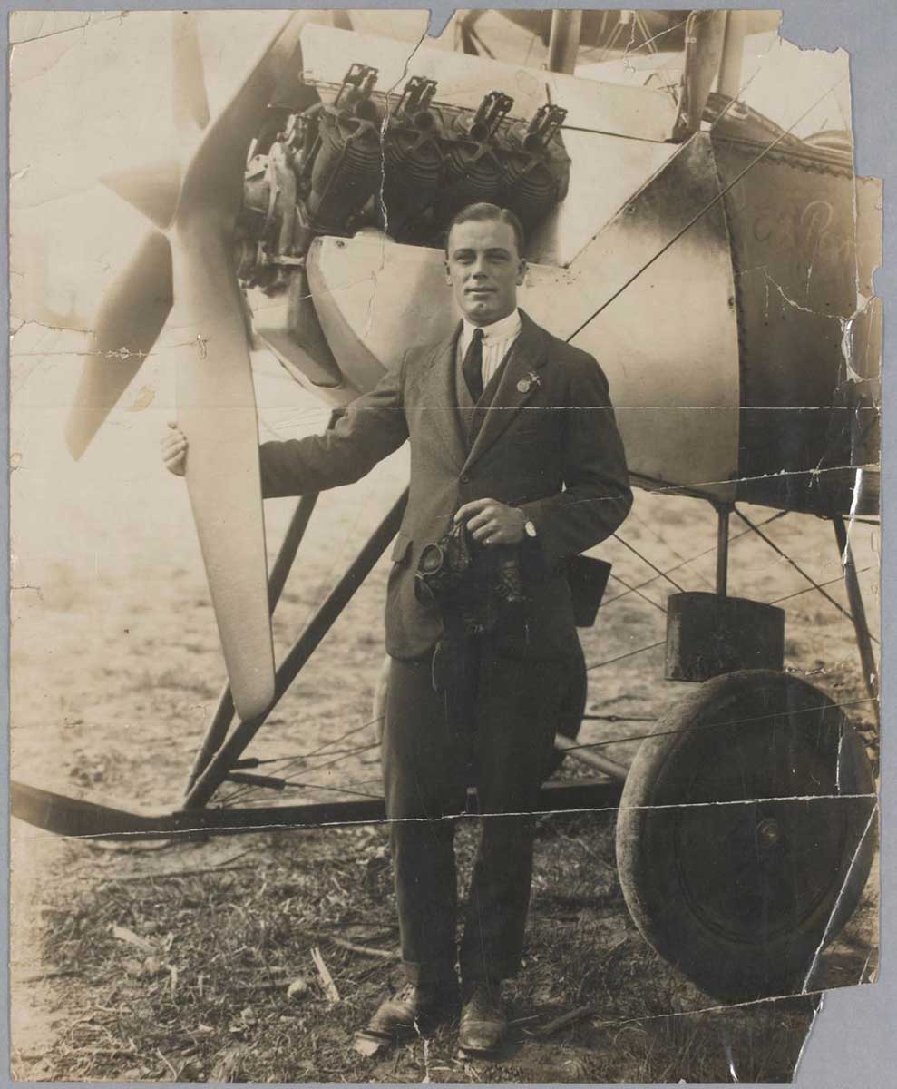 A black and white photo of a man standing at the front of plane, with one hand resting on a propeller blade. - click to view larger image