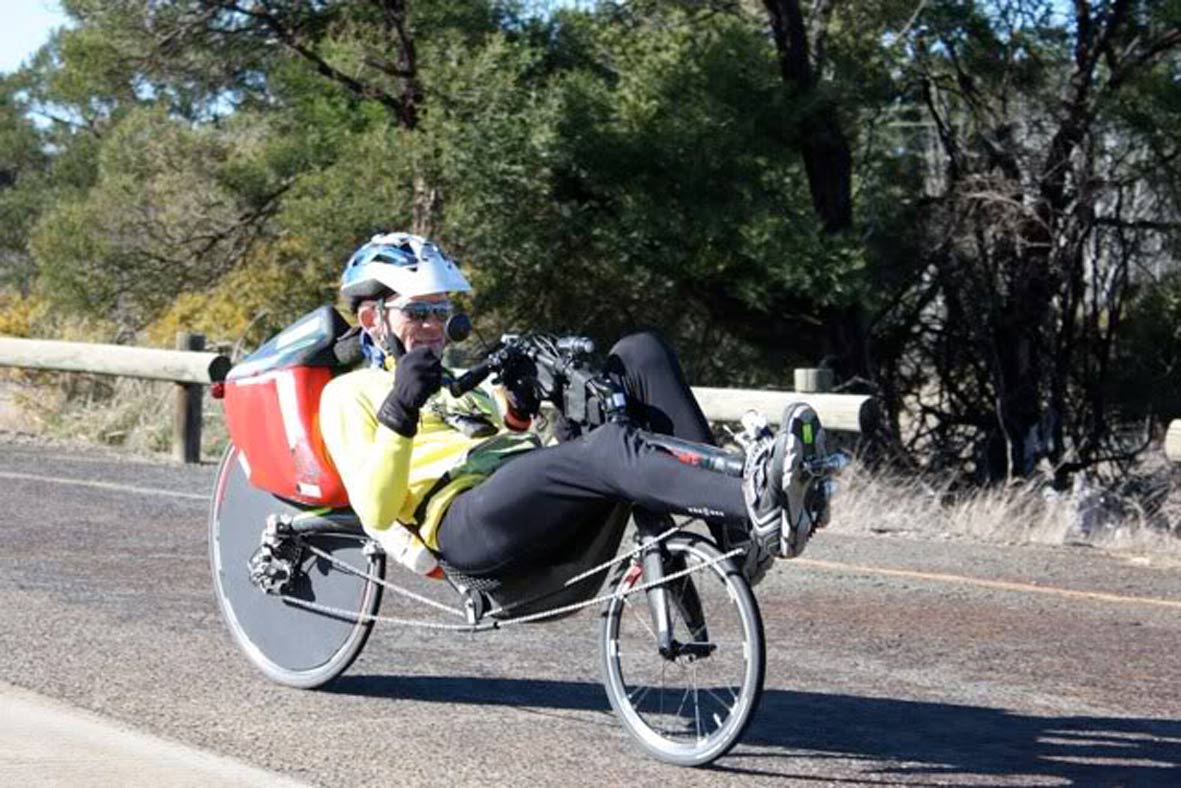 Colour photograph showing a cyclist on a recumbent bike, on a bitumen road. The rider is looking ahead and has made the 'thumbs up' sign with his right hand. - click to view larger image
