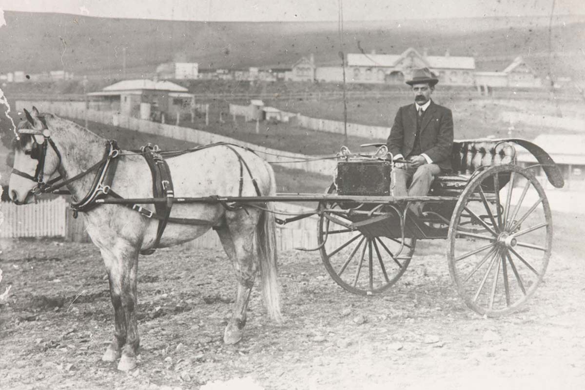 Black and white photo of a man sitting in a sulky hitched to a horse.