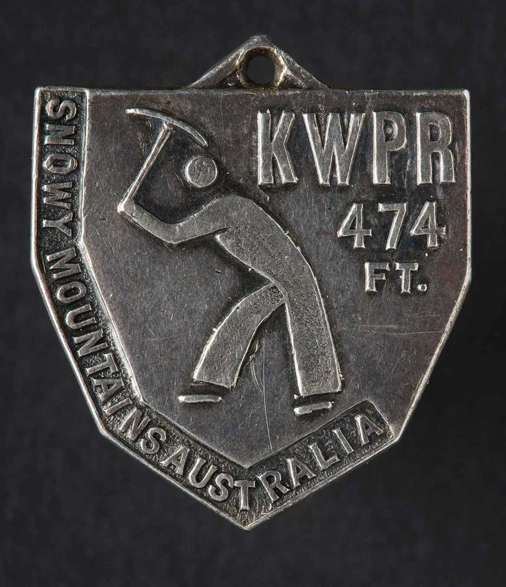 An image of a worker holding a tool above his head and in a digging stance is in the centre of the medal. 'KWPR 474 FT' is written in the top right corner and 'Snowy Mountains Australia' is written down the left hand side. - click to view larger image