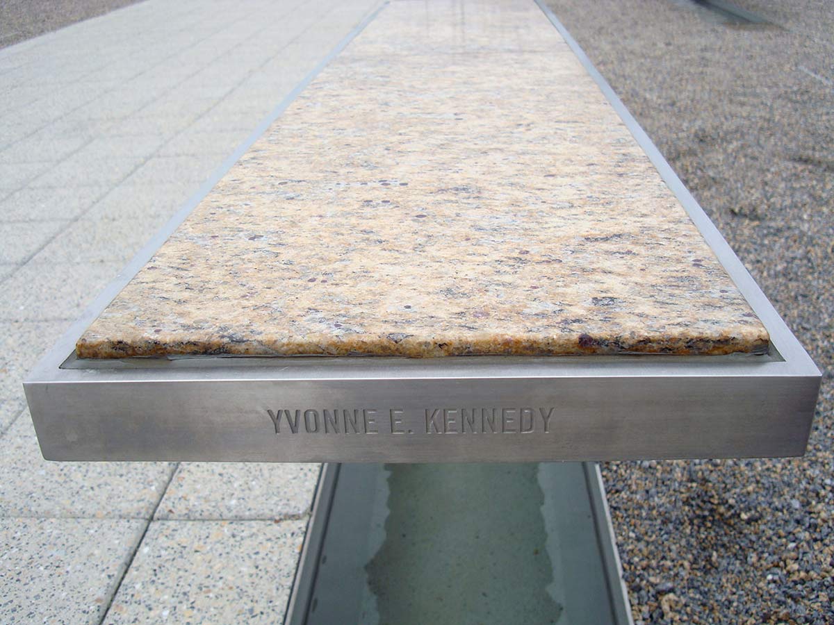 Far end of a silver rectangular metallic bench with a light marble top. 'YVONNE E. KENNEDY' is engraved in the side of the bench, which sits abover a pool of water. - click to view larger image