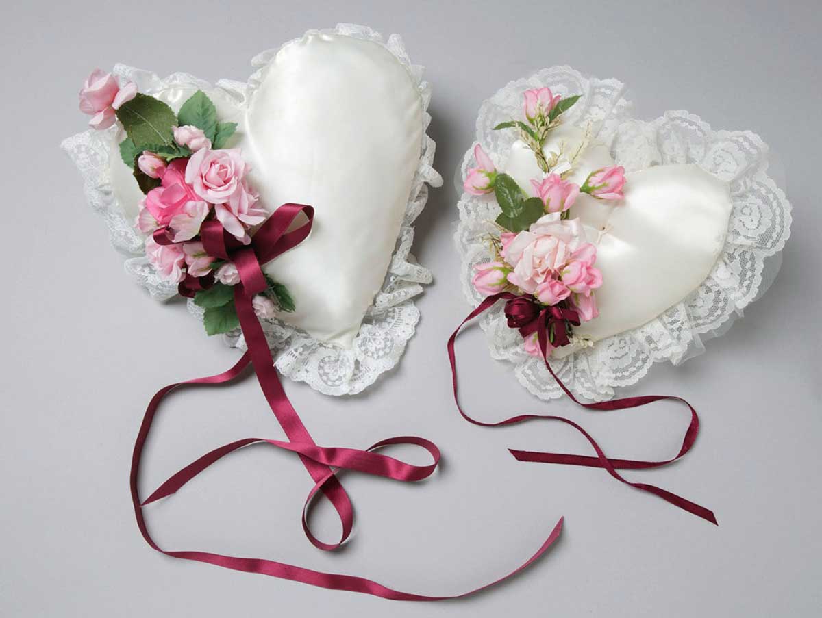 Two love-heart shaped cushions, similar in appearance, decorated with maroon ribbons and pink flowers. - click to view larger image