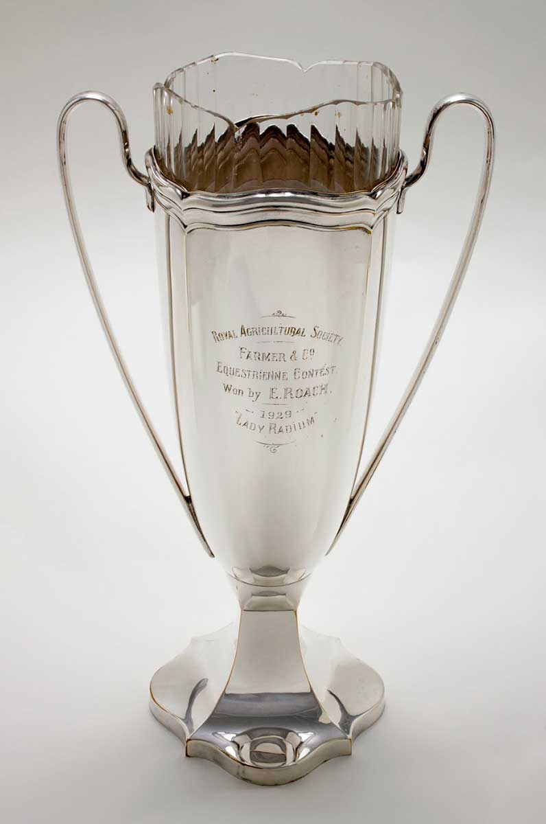 A large silver plate presentation trophy cup. The cup features a roughly square shaped base with scalloped edges tapering up into a square cross section at the immediate bottom of the cup. This area then tapers out into the tall, roughly cylindrical shaped bowl of the cup. The lip of the cup is scalloped featuring decorative ridges around its circumference. Two long decorative handles run either side of the cup down most of its length. Engraved on a central panel of the bowl of the cup is the text 'ROYAL AGRICULTURAL SOCIETY / FARMER & CO. / EQUESTRIENNE CONTEST / Won by E. ROACH, / 1929 / 'LADY RADIUM'. - click to view larger image