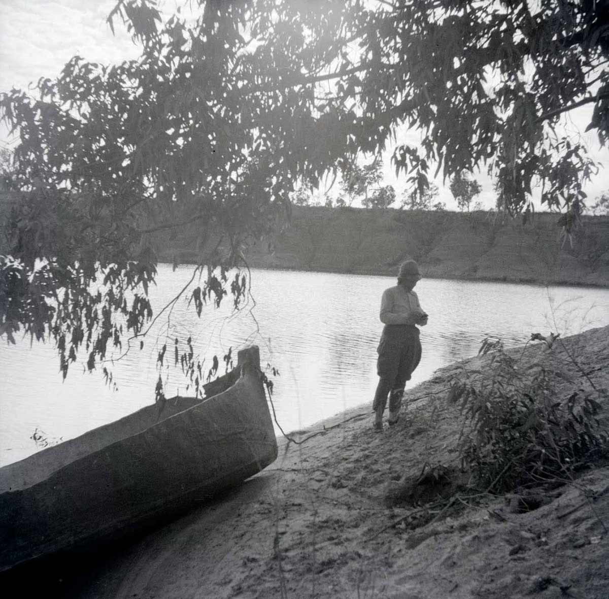 A black and white photographic negative that depicts an adult standing on a riverbank next to a canoe. - click to view larger image