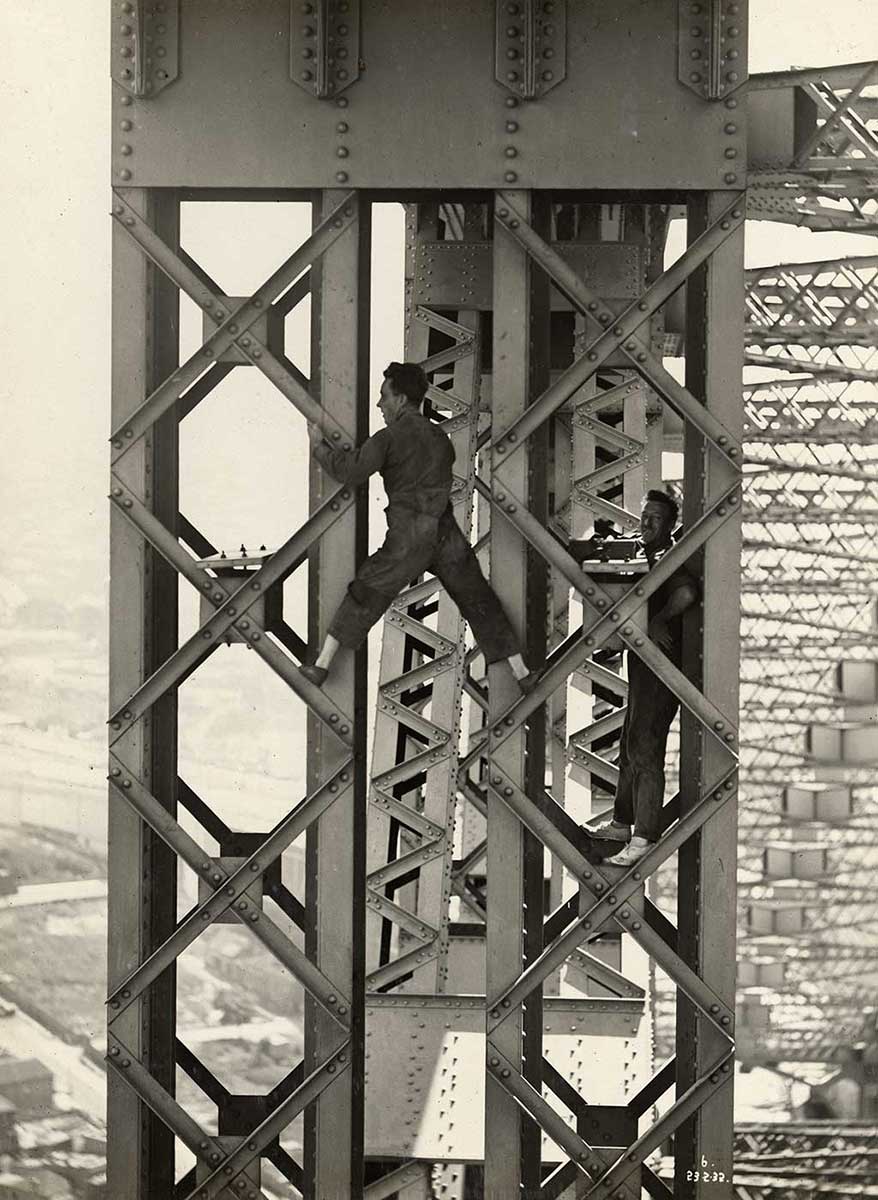 A man stands astride two sections of metal bridgework. He wars overalls and shoes without socks. Another man stands smiling while resting on another section of bridge at right. - click to view larger image