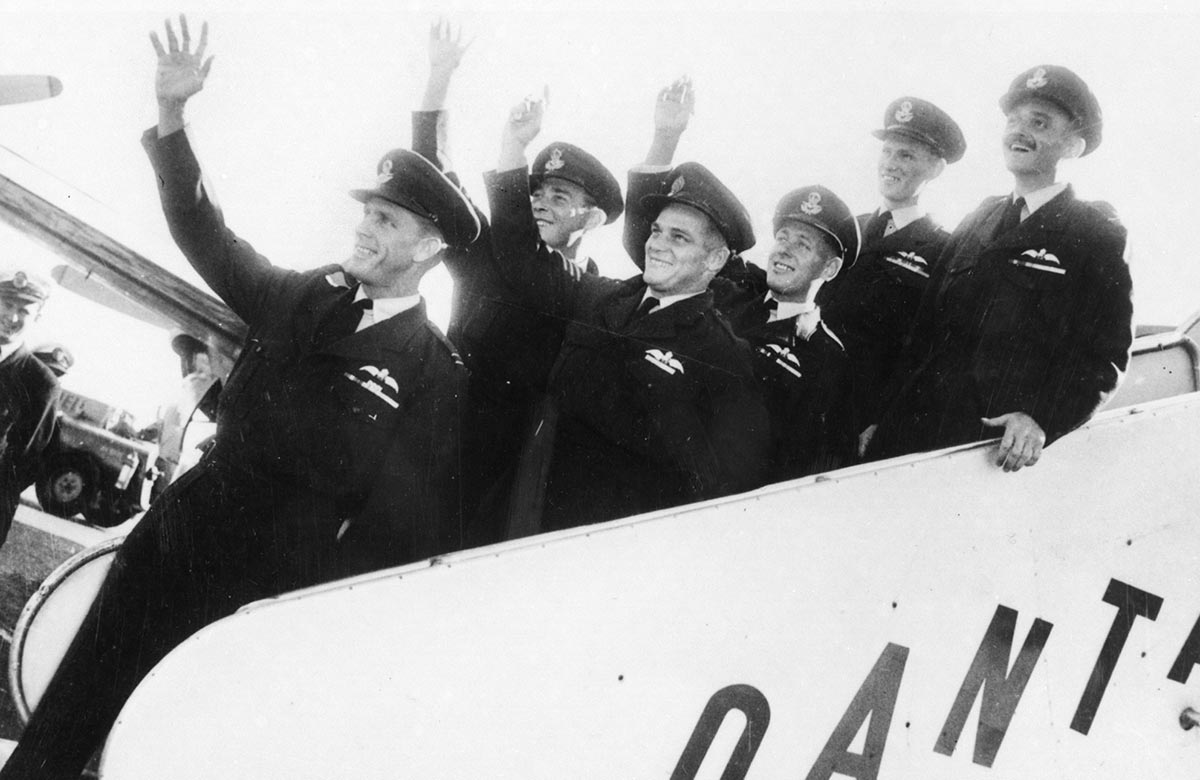Six men in RAAF uniform smiling and waving while standing on the moveable staircase attached to a plane