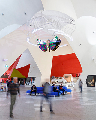 Image showing the interior of a building with people moving through the space and a colourful moth sculpture hanging from the ceiling. 