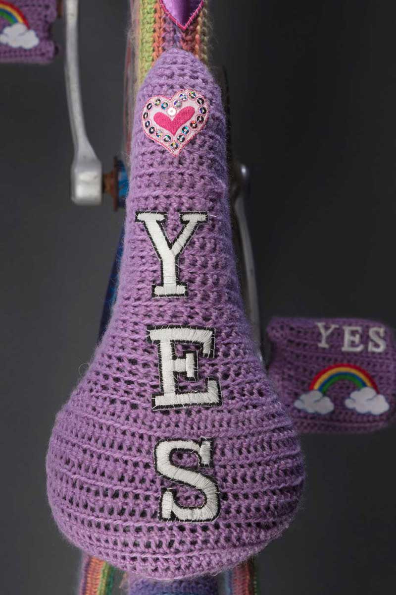 Bike seat covered in mauve wool with the word 'YES' woven vertically beneath a red heart. - click to view larger image