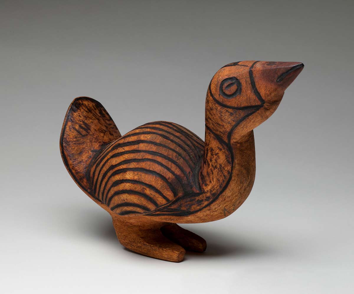 Black and brown wooden bird sculpture standing upright on two legs with the neck straight up and the head looking upwards and an upright, short broad tail. The top exterior of the carved bird has burnt pokerwork designs around the eyes, beak and nostrils with the head, neck and tail covered in a lightly shaded speckled pattern and the body with parallel curved lines. - click to view larger image