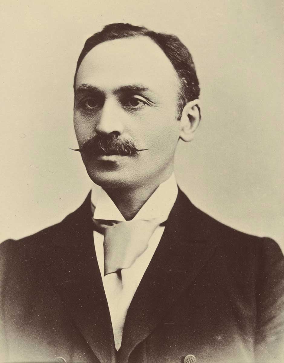 studio portrait of young man, unsmiling with pointy moustache and cravat - click to view larger image