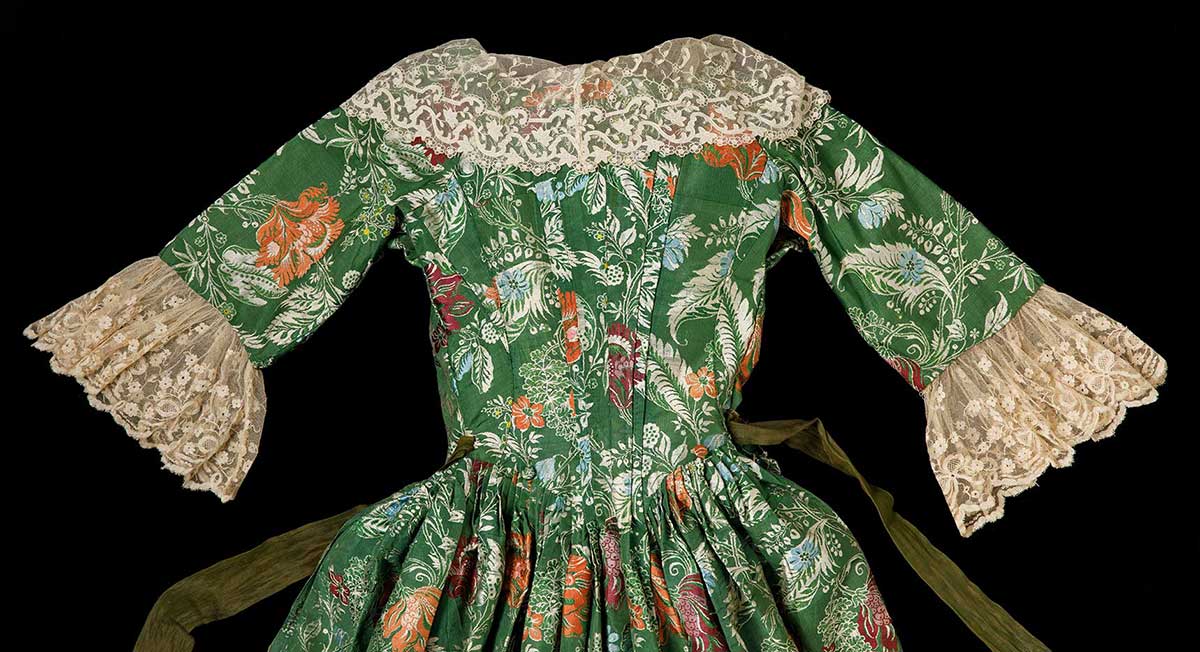 A detail image of the top part of the back of a green and floral dress with lace trim on the sleeves and collar with a long green sash like belt stitched at the waist. - click to view larger image
