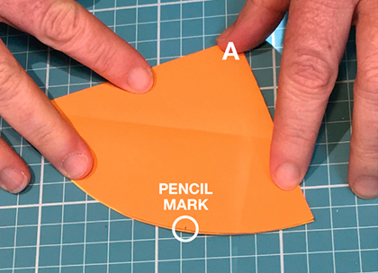 Fingers hold down a piece of orange paper. The letter B in white font is superimposed on the top left corner of the paper and the capital letter A (also in white font) is superimposed on a pointy end in the centre. A few centimetres to the left is a small block of black text in capital letters which reads 'curved edge' with an arrow pointing to the curved edge of the paper.