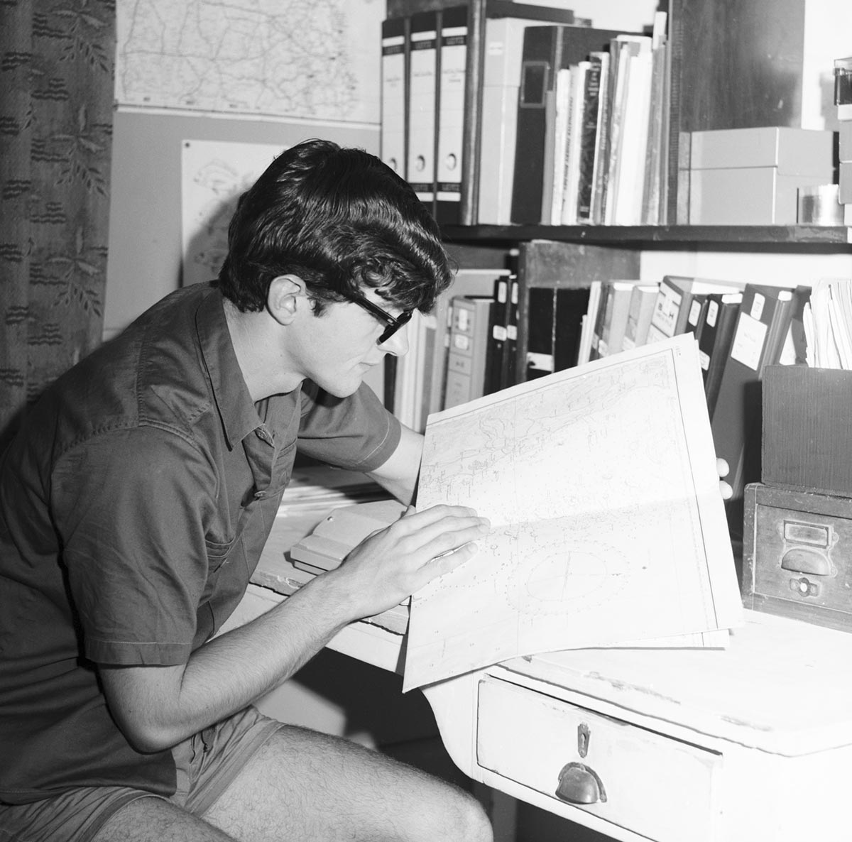 Young man with glasses seated at desk examining large book, possibly an atlas. - click to view larger image