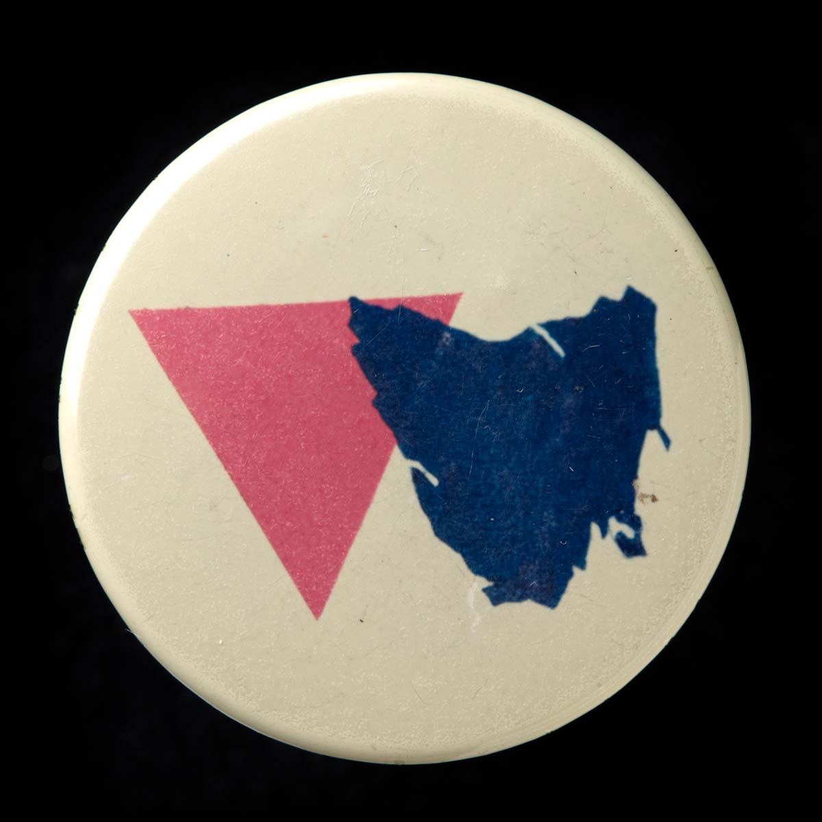 A political badge with a cream background and a design which features a pink triangle and a map of Tasmania in navy blue. - click to view larger image