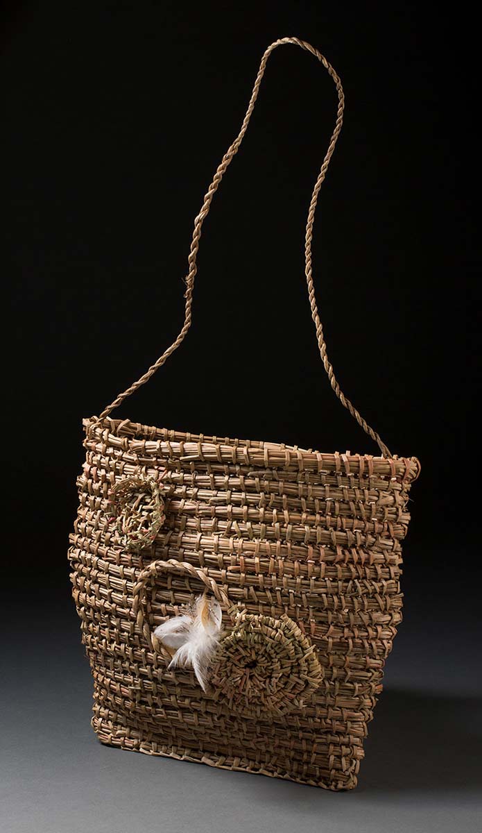 A woven bag that is almost square in shape and has a long plaited handle. The front side of the bag has woven decorative coiled circles with white and brown feathers attached. - click to view larger image