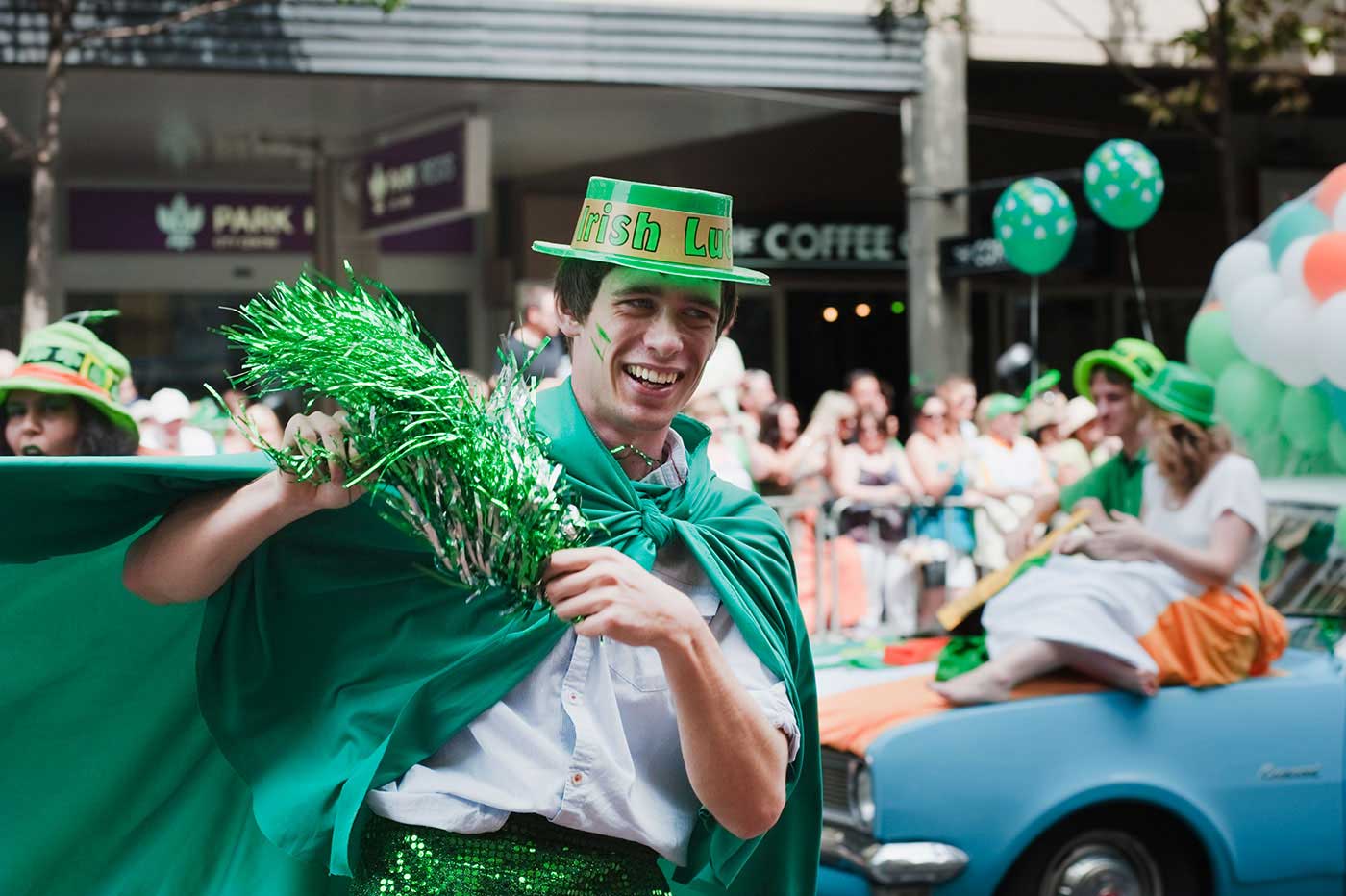 A man is wearing a green costume with a gimmick hat surrounded by other people wearing green in a procession on a city street. - click to view larger image