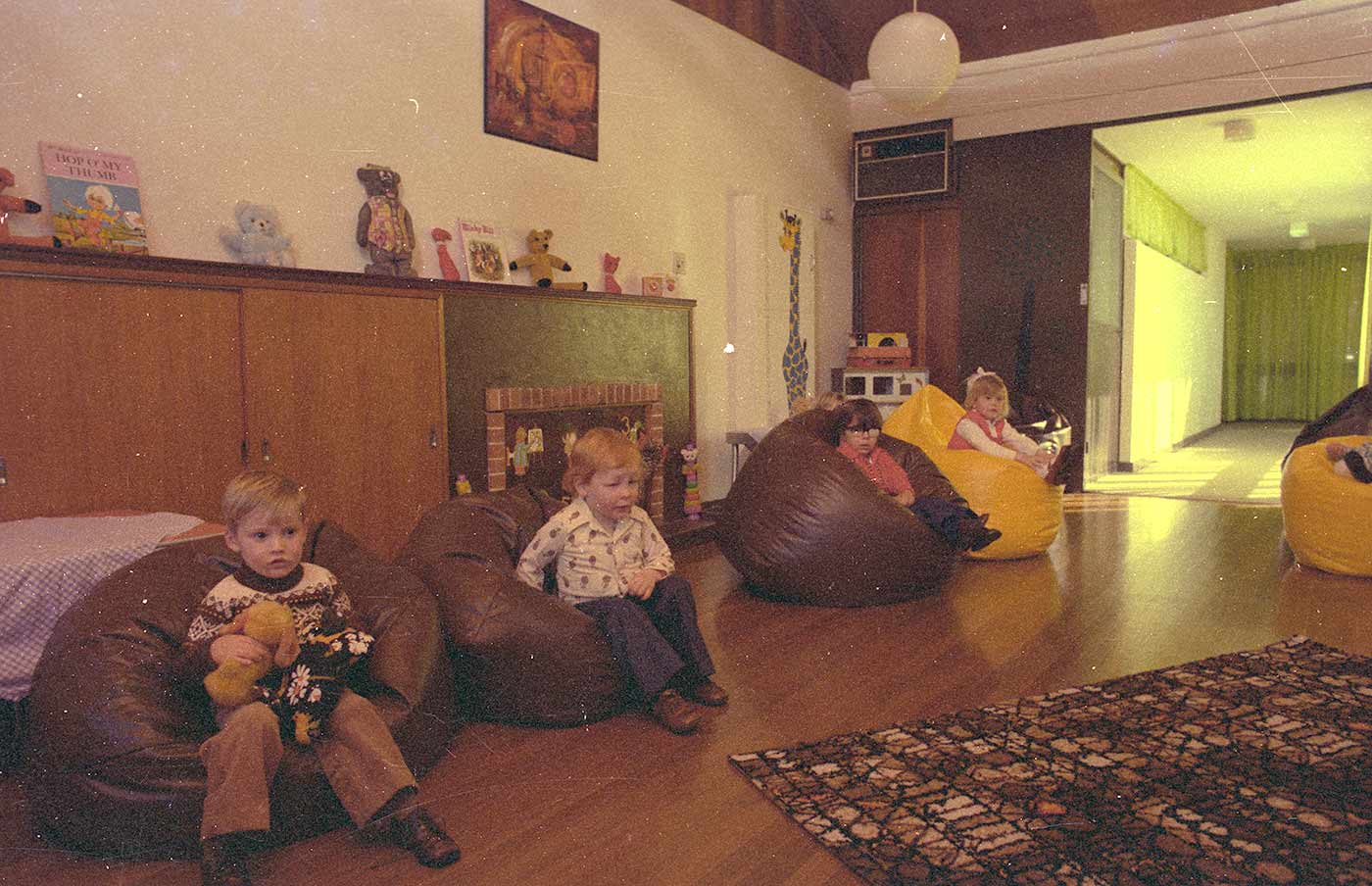 Colour photograph showing four small children sitting on bean bags. A fifth is partially visible at right. The child in the foreground holds a soft toy. Toys and books sit atop cupboards and above a fireplace at the rear. - click to view larger image
