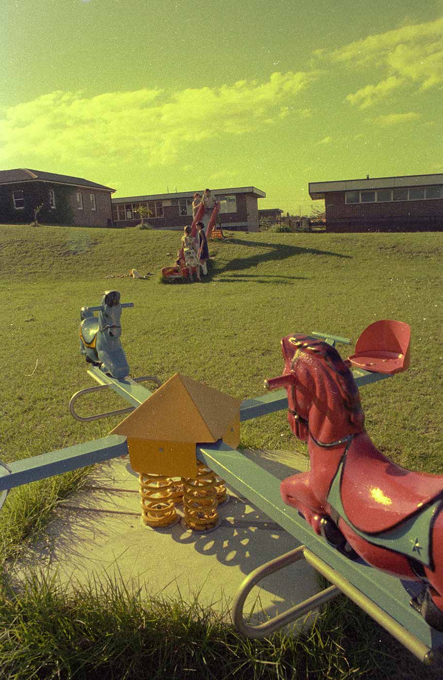 Colour photograph showing a four-ended seesaw with horses on one bar, and seats on the other. Children play in the distance on a slippery dip at the top of a grassed hill. Several brick buildings are visible in the background. - click to view larger image