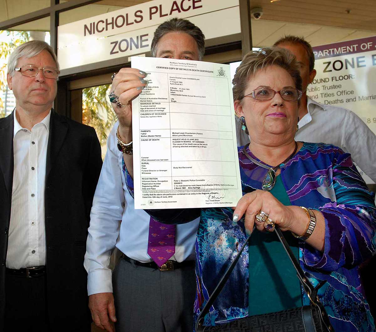 A woman standing outside of an office building is holding up an official document. A group of men in business suits stand behind her.