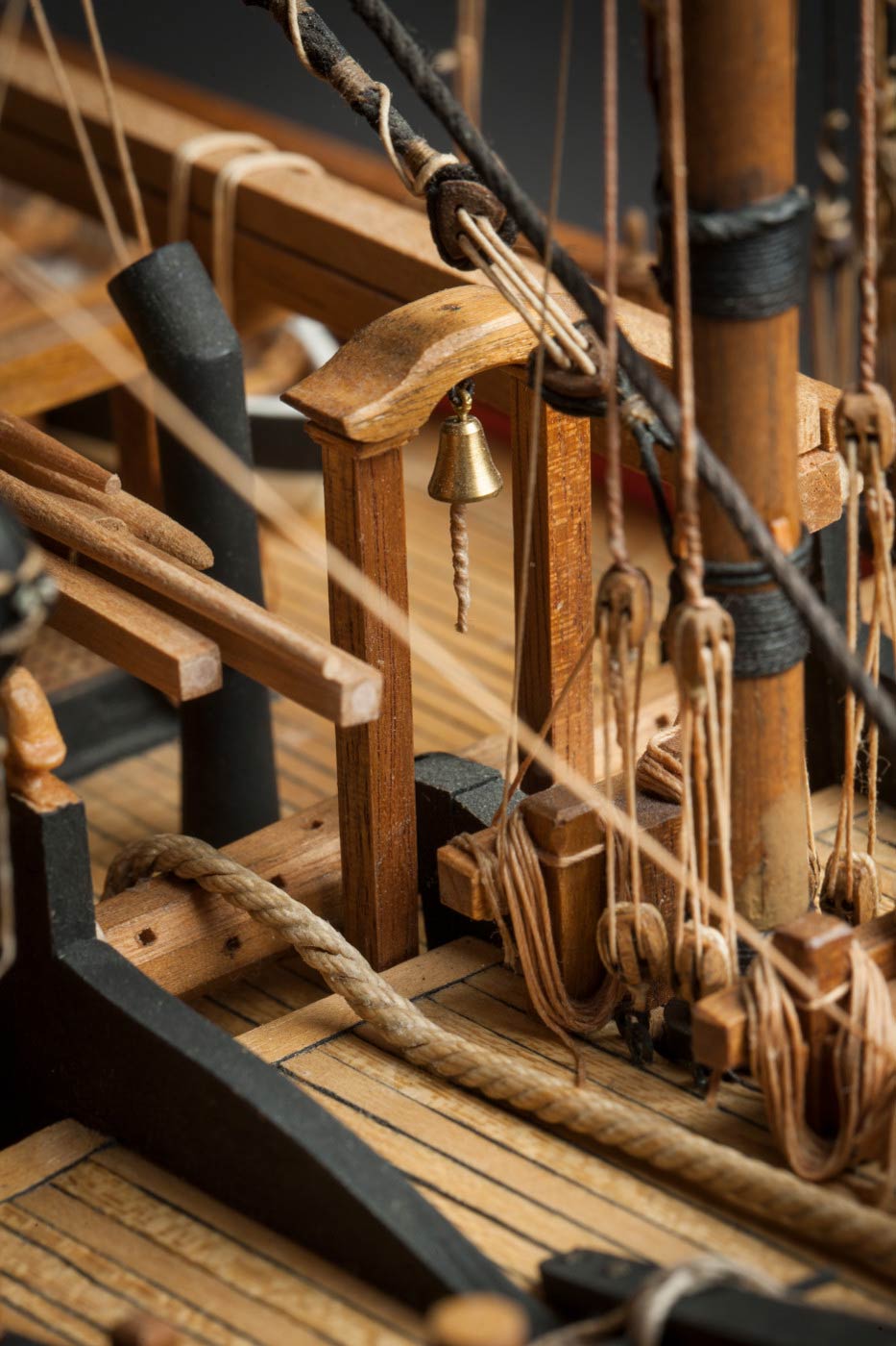 Detail image showing a small, gold-coloured bell, suspending among the rigging of a wooden sailing ship. - click to view larger image