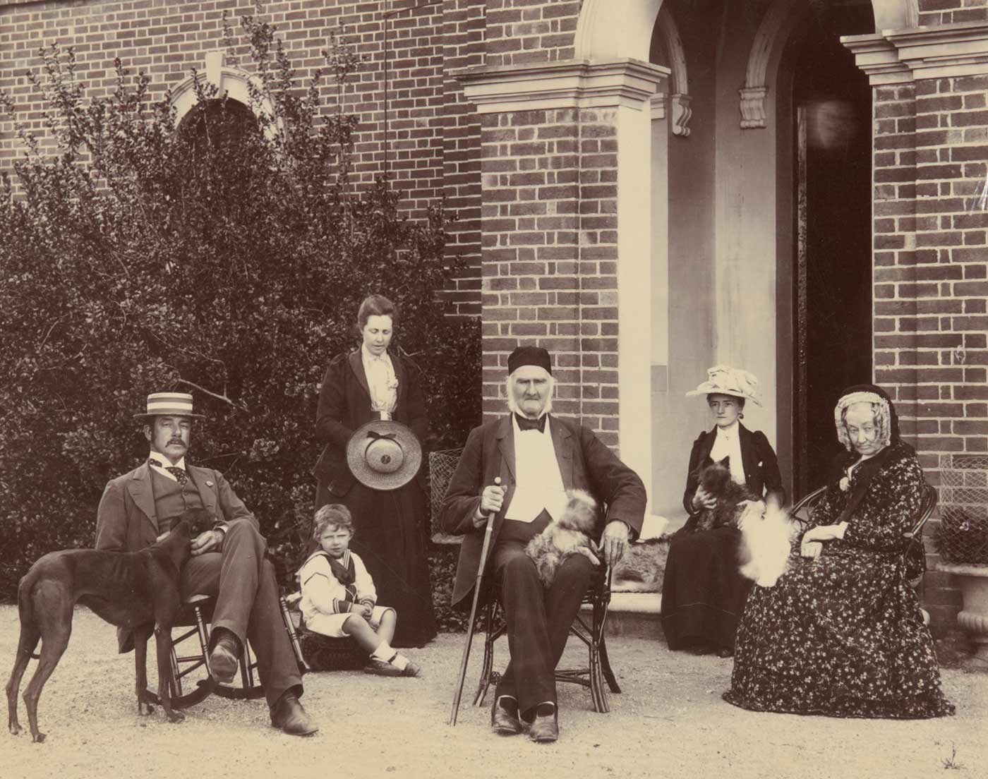 Black and white photo showing two men, three women and a child outside a brick building. The man at centre holds a walking stick and has a small dog sitting on this lap. The other man sits in a rocking chair, with the head of a kangaroo dog resting on his lap. To his right is a small boy in a sailor suit, and a woman, standing, who is looking down. The women seated, far right, both hav animals resting on their laps. - click to view larger image