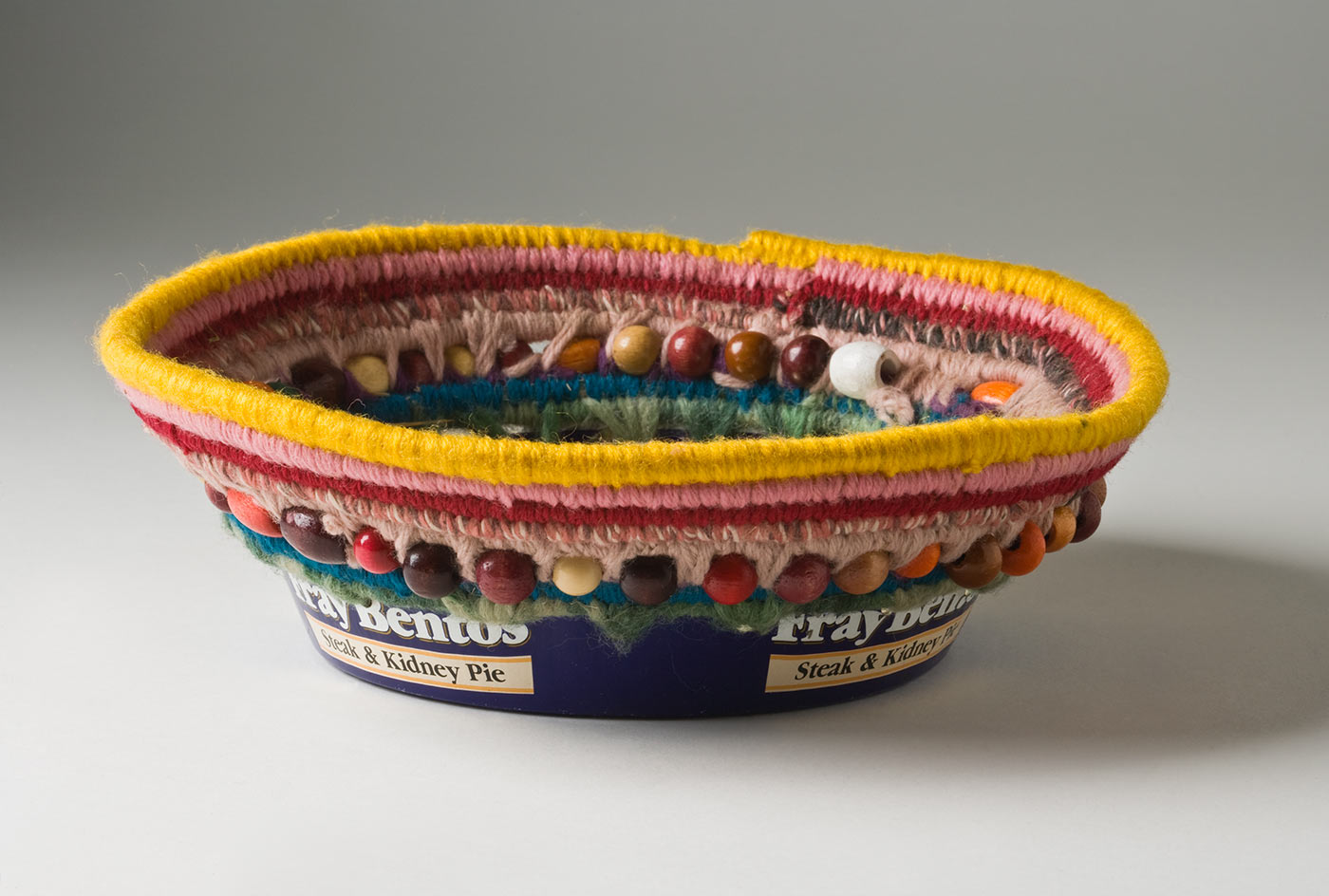A circular coiled striped yarn basket over plant fibre with bead decoration, which opens out towards the top and has a metal base. The base of the basket is a blue and white coloured metal tin with burn marks to the bottom which includes the text 'Fray Bentos / Steak and Kidney Pie'. Yarn is used to attach the base to the top section through holes that have been punched in the tin. The basket has a stripe of yellow at the top edge followed by pink, dark pink, brown and mid pink, peach, teal and light green. Multicoloured oval and round wood beads are attached within the yarn coils at the top of the metal base. - click to view larger image