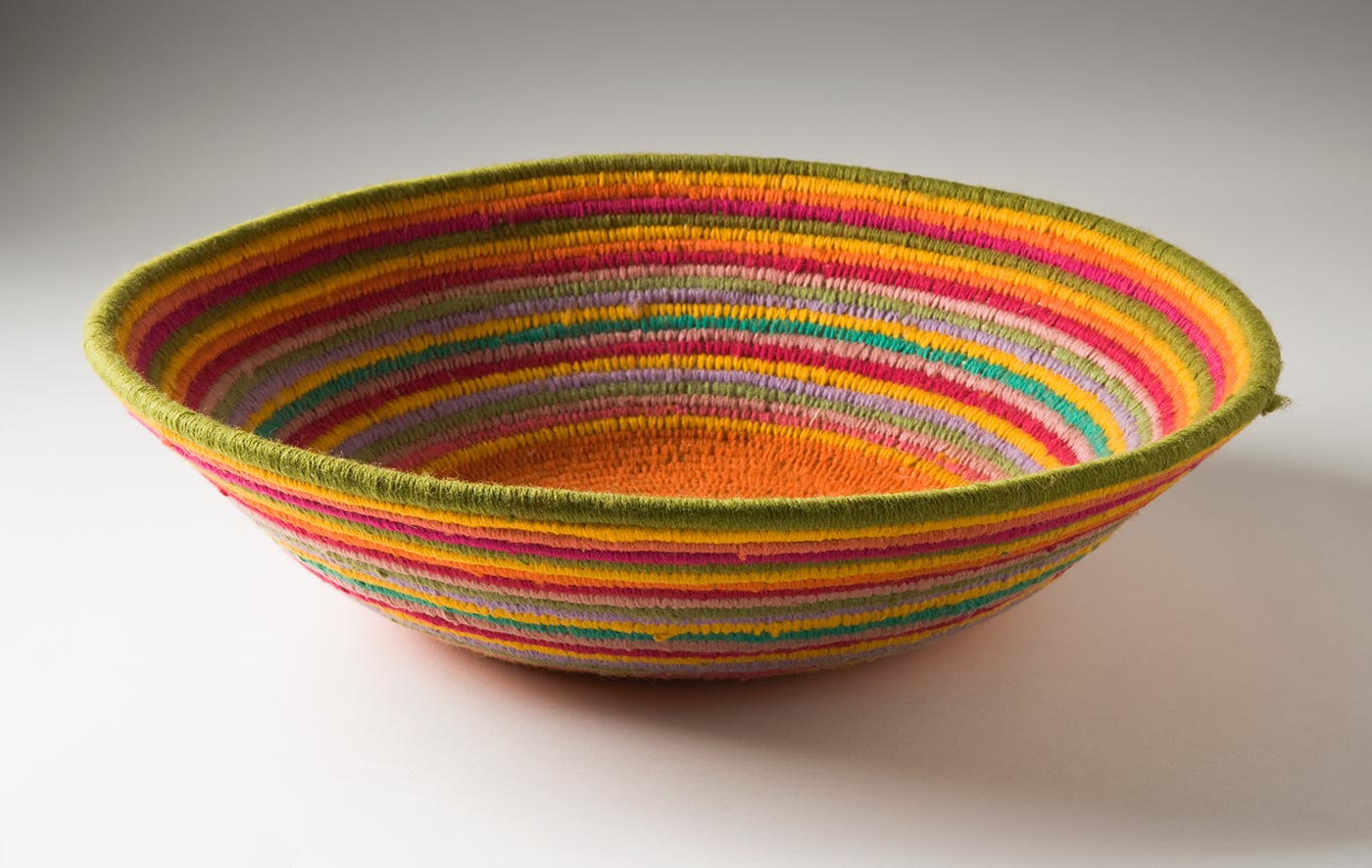A multi-coloured circular coiled bowl-shaped basket made of yarn and plant fibre. The centre of the basket is in orange yarn which persists out to where the edges start to turn up. The rest of the basket is in horizontal stripes of yellow, dark pink, light pink, lime green and lavender yarn. - click to view larger image