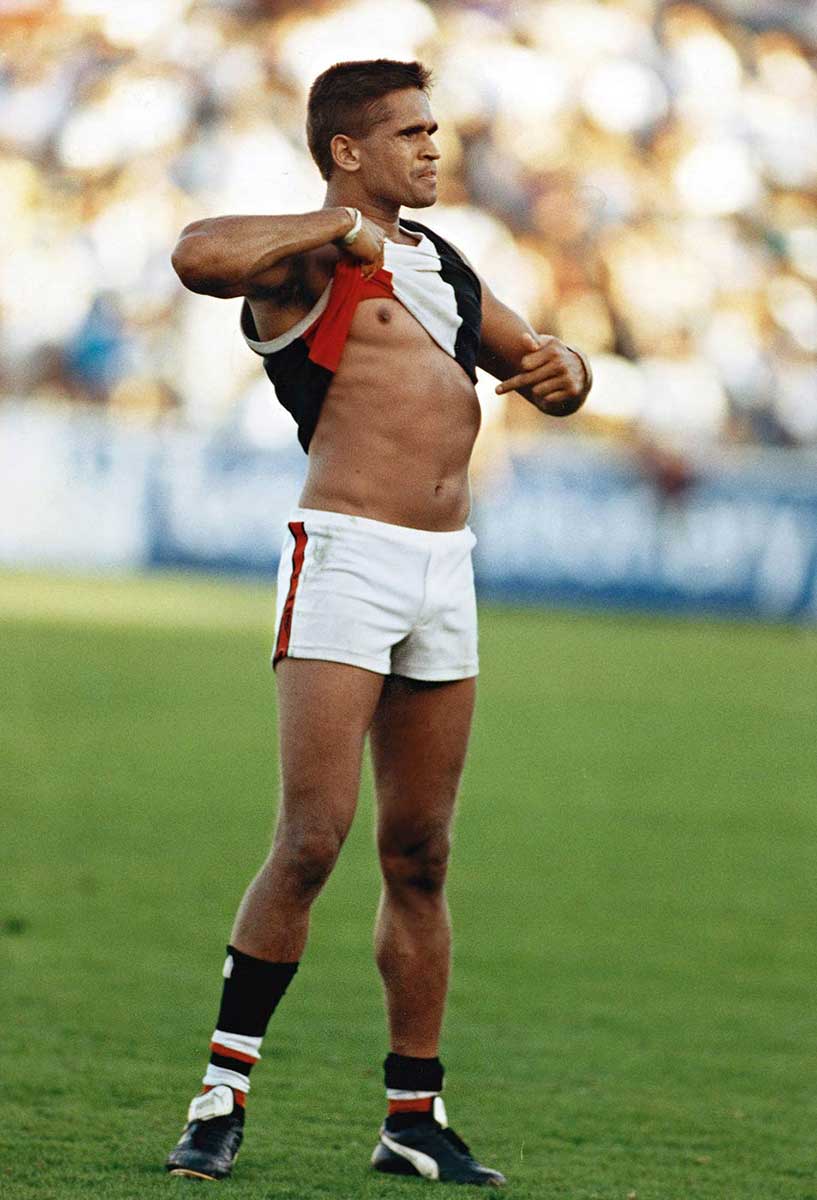 Full length colour photo of an Aboriginal AFL player standing on the field, lifting up his jumper with one hand and pointing at his belly with the other. He has a determined and defiant expression on his face. - click to view larger image