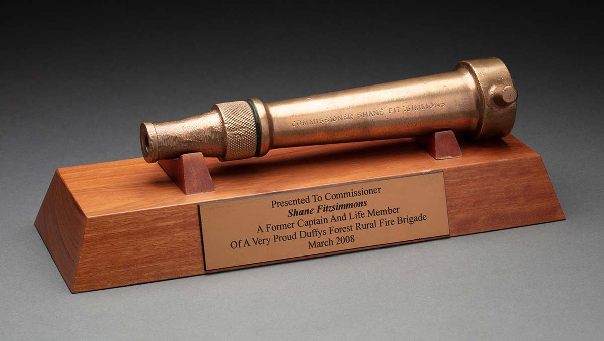 A trophy which features a brass nozzle from a fire hose engraved with the text ‘COMMISSIONER SHANE FITZSIMMONS’ mounted on a wooden base. A plaque fixed to one side is printed with the text: ‘PRESENTED TO COMMISSIONER SHANE FITZSIMMONS / A FORMER CAPTAIN AND LIFE MEMBER / OF A VERY PROUD DUFFYS FOREST RURAL FIRE BRIGADE / MARCH 2008.