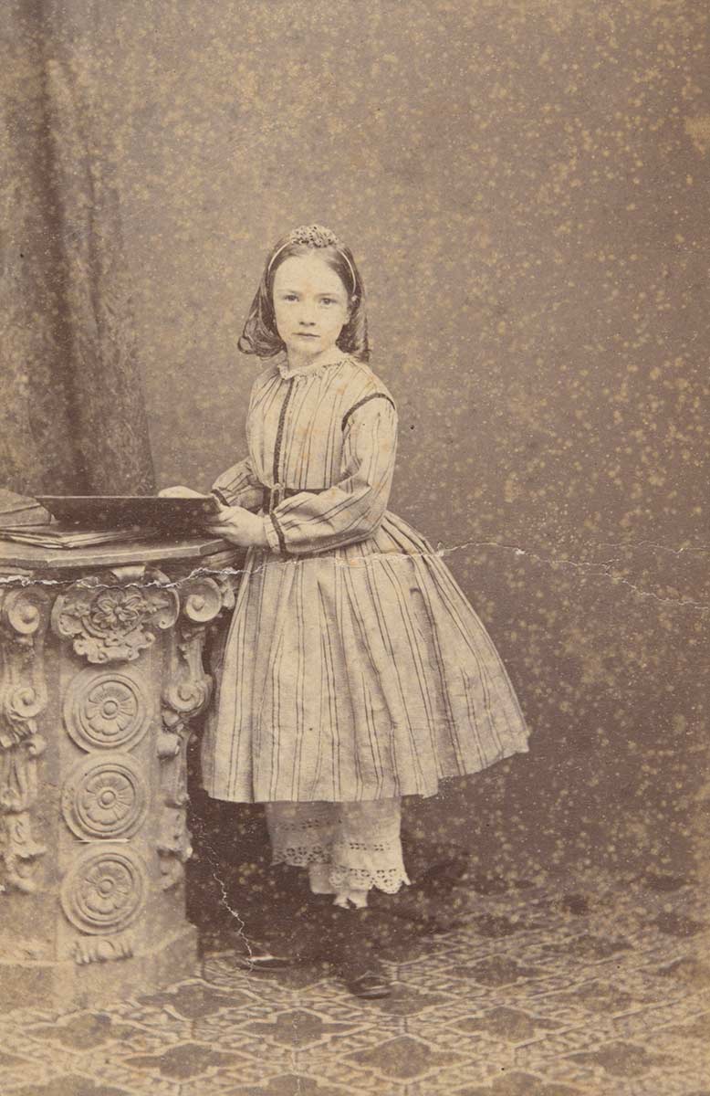 A small photograph of Lilian Faithfull as a young girl. She is standing beside an ornate short column on which a book is placed. - click to view larger image