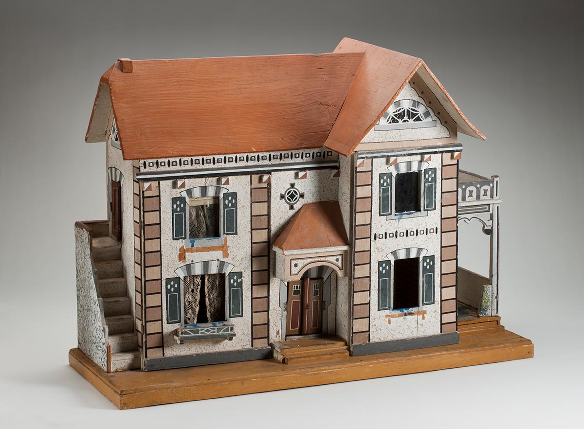 A double storey dolls house with two panels at the front of the house that open to reveal the interior. It has a pitched brown roof, a set of stairs leading up to a door on the second floor on the left, a covered front entrance in the middle, and a covered porch on the right. It also includes lace curtains in top and bottom windows on the left panel. The exterior is decorated with painted gables, window coverings and stone effects above the windows and on the corners of the house. The double door main entrance on the front is finished with painted wooden strips representing lintels, sills, and architraves, and can be opened and closed. The house is secured to a wooden base. - click to view larger image