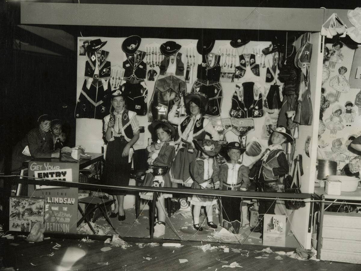 Black and white photograph of a group of people in costume standing in a temporary shop-front with costumes hanging on the wall. - click to view larger image