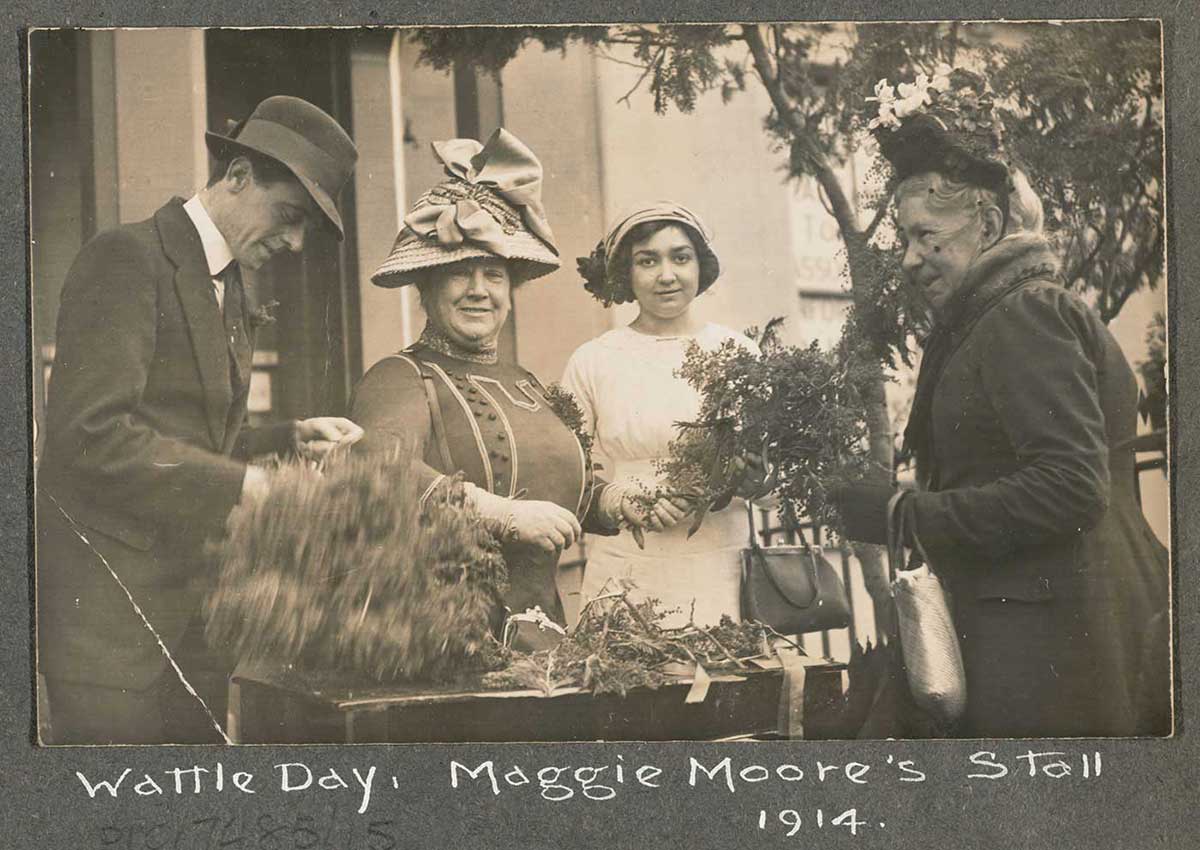 Photograph mounted on board, showing three women and a man standing in front of table loaded with wattle sprays. 'Wattle Day. Maggie Moore's Stall 1914' is handwritten across the bottom of the board. - click to view larger image