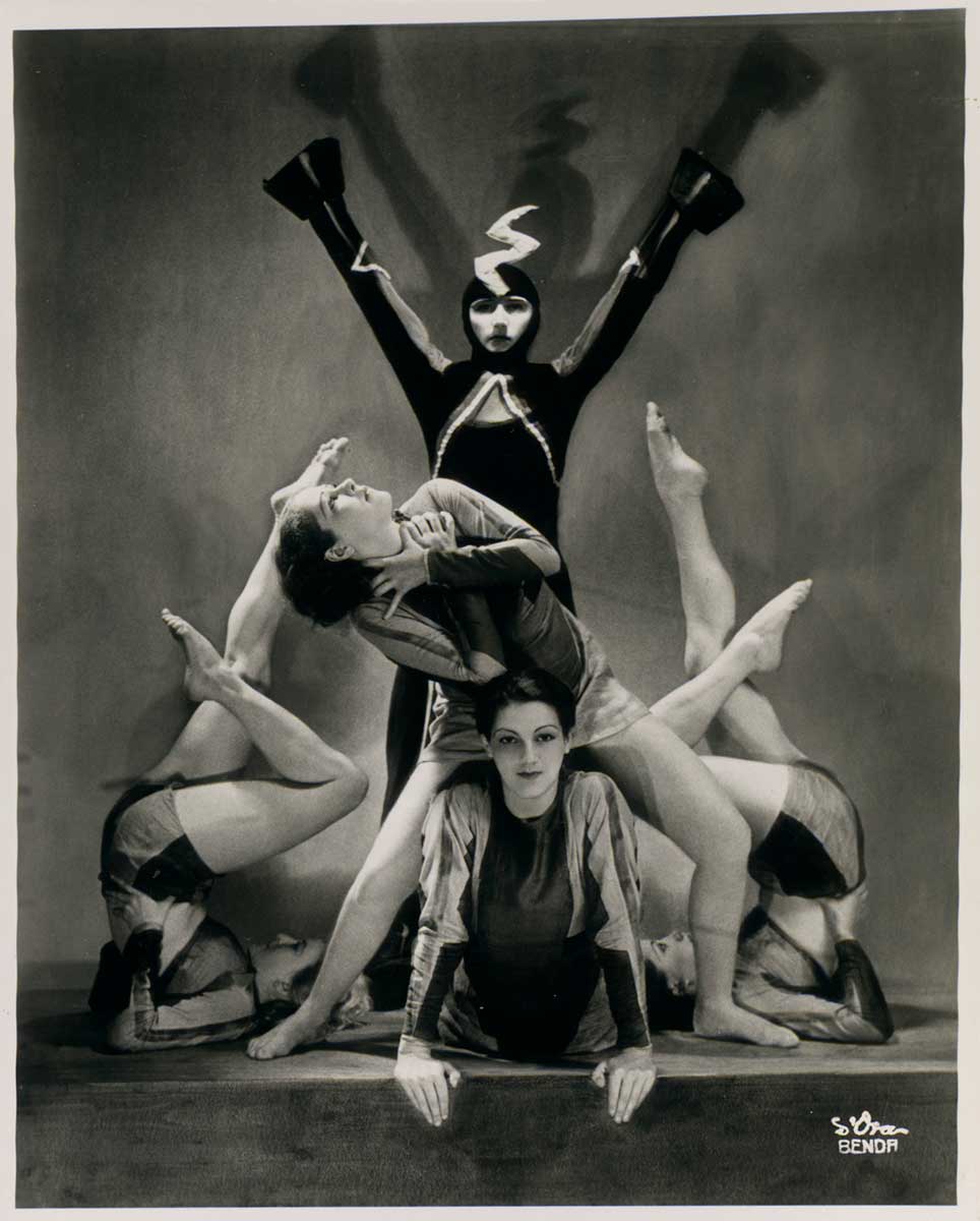 Black-and-white photograph of five dancers in poses from a ‘Demon Machine’ performance. A dancer sits in the middle of the stage, propped up on her hands with her legs behind her and looking towards the camera. Another dancer stands above her with her legs on either side of the first dancer’s body. The second dancer has her hands clutched at her neck and looks upwards. On either side of these two dancers are two more, lying on their upper backs with their hips and legs pointed into the air. Behind all these dancers is the fifth, wearing a costume meant to signify ‘the machine’ in the performance. She stands with her arms raised into the air and her costume is dark with sharp lines and a lightning-bolt shaped headpiece. - click to view larger image