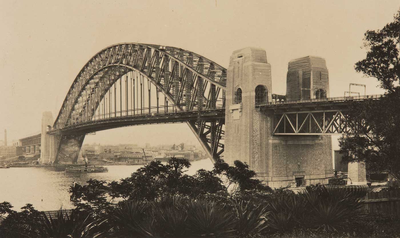 A black and white photograph of the Sydney Harbour Bridge, with a steam ferry passing under it. - click to view larger image