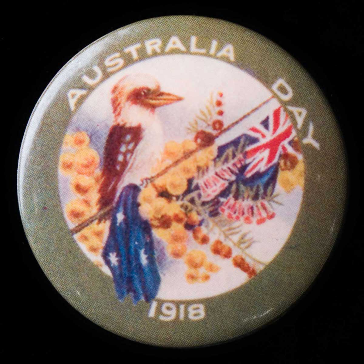 Circular badge with an olive-coloured border with the words 'Australia Day 1918' printed in white. The central image shows a kookaburra sitting on an Australian flag, surrounded by yellow wattle blossoms and red trumpet-like flowers. - click to view larger image