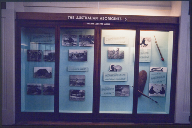 Museum cabinet displaying photos and First Nations objects. The cabinet has four glass front panels, each framed with timber. The panels are tall and rectangular. Above the glass panels on the outside timber cabinet frame is a sign in raised letters that says 'The Australian Aborigines 5'. Beneath that in smaller raised letters is 'Shelters and fire-making'. In the cabinet are photographs, text panels and three objects. The photographs and most of the text panels are grouped in the centre and left of the cabinet. Two text panels with images and the three objects are grouped at the right end of the cabinet. The objects appear to be a wooden shield, a spear thrower and a stick tool. A single fluorescent light is above the objects in the right side of the cabinet. The photographs and text panels in the left side of the cabinet are in shadow and less easy to see. - click to view larger image