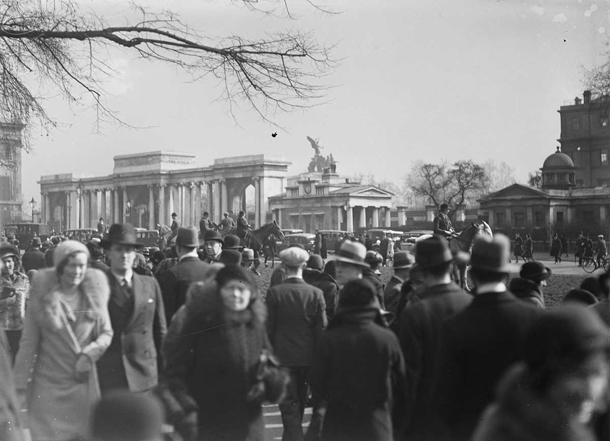 Crowd in Hyde Park, London, England 1931 or 1932. The crowd in the photograph appears to be all men and women. Many of the men wear long coats over suits, as well as hats or soft caps. The women in the photograph wear coats and hats as well. Much of the crowd is in the foreground and middle ground. In the background can be seen men on horseback. Beyond them are various buildings and a monumental facade. The cloudless sky is visible above the buildings and the facade. The bare branch of a tree projects into the photograph in the top left corner.