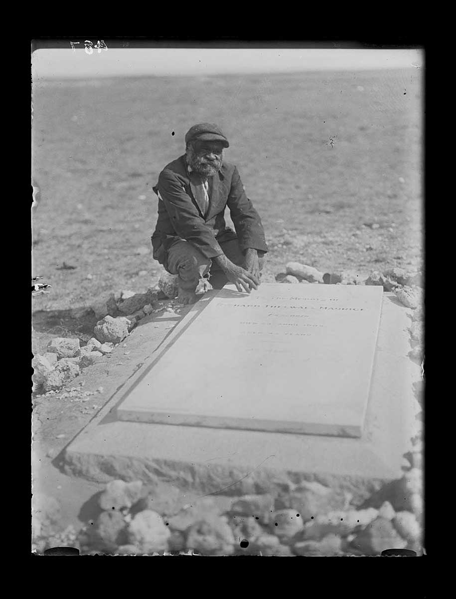 An Aboriginal man at a European grave in a remote location, Yalata Station, South Australia 1920. The grave is marked by a large horizontal slab; on top of that is a plaque with information of the deceased. The Aboriginal man squats at the top left corner of the grave, with his hands almost touching the plaque. He wears a coat, trousers and peaked cap. The background is open ground leading to the horizon. - click to view larger image
