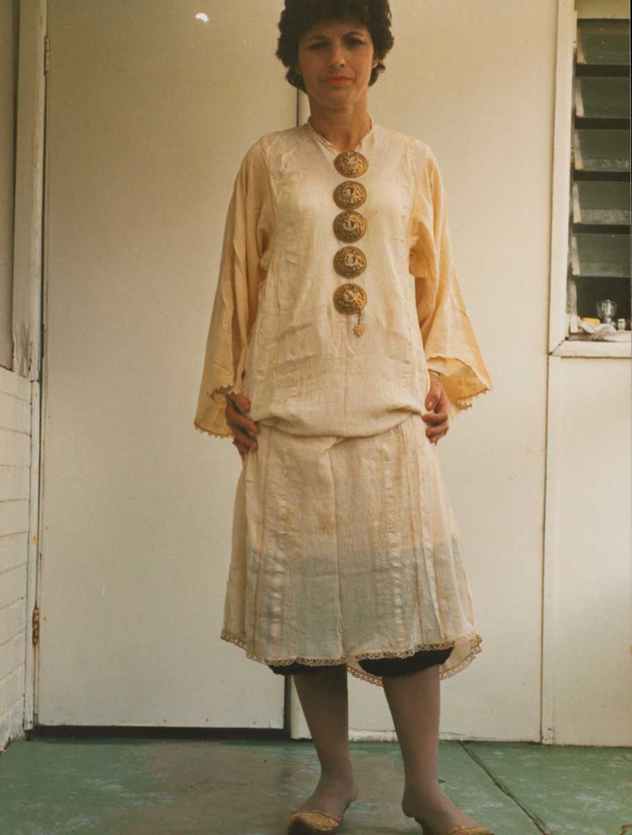 Portrait photograph of a woman with short dark hair wearing a knee-length off-white chemise with long, loose sleeves. Under the chemise are a pair of knee-length black and cream pantaloons, only just visible under the chemise. The woman wears five gold button brooches, running down the chest of the chemise, and a pair of embroidered gold slippers with a small heel. She stands facing the camera with her hands by her side. - click to view larger image