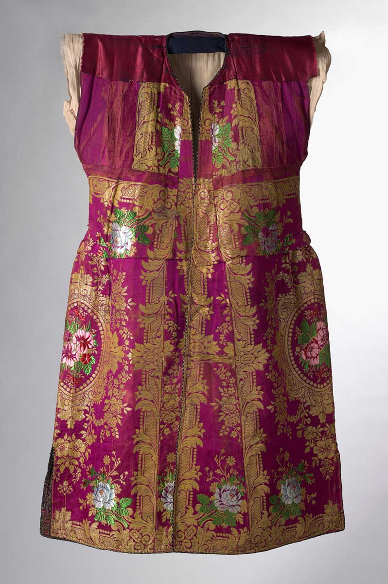 Crimson full-length tunic, part of Castellorizian formal attire worn by Rose Pappas Description Greek crimson full-length tunic with a rounded plain neckline and two side splits. It is fully lined with highly decorative damask weave fabric at the front opening and around the hem. It has orange silk to the main body. The main fabric is of highly decorative damask weave (composite). Decorated with black and gold braid, embroidered floral motifs in gold thread, and various coloured silk threads. Tuck sewn at waist. The bodice section is lined with calico. - click to view larger image